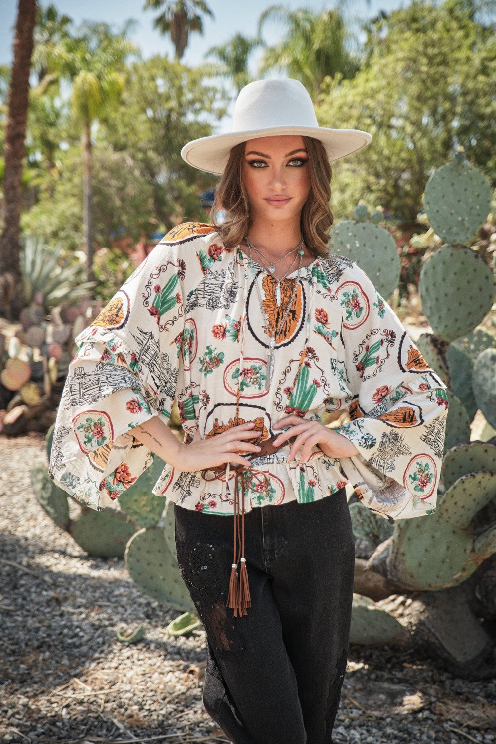 Woman wearing a patterned blouse and white hat in a desert setting with cacti.